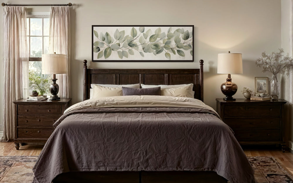 Long horizontal framed canvas wall art of eucalyptus leaves on rustic wood above a bed.