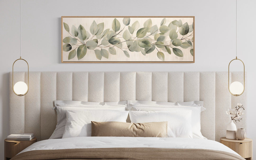 Long horizontal framed canvas wall art of eucalyptus leaves hanging above a bed in a bedroom.