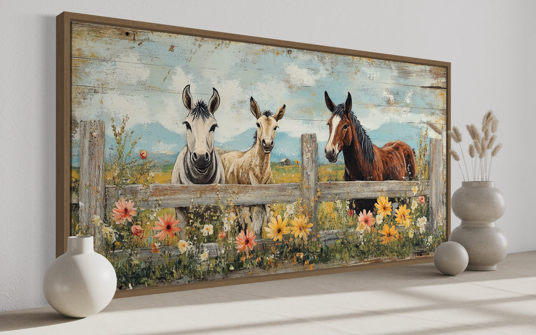 Framed canvas wall art of two donkeys and a horse behind a rustic fence with colorful wildflowers.