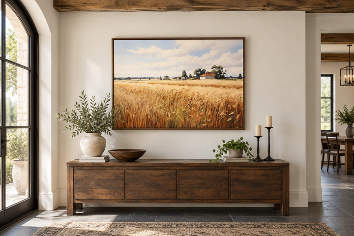 Rustic wheat field landscape painting in a wood frame above a dark wood sideboard.