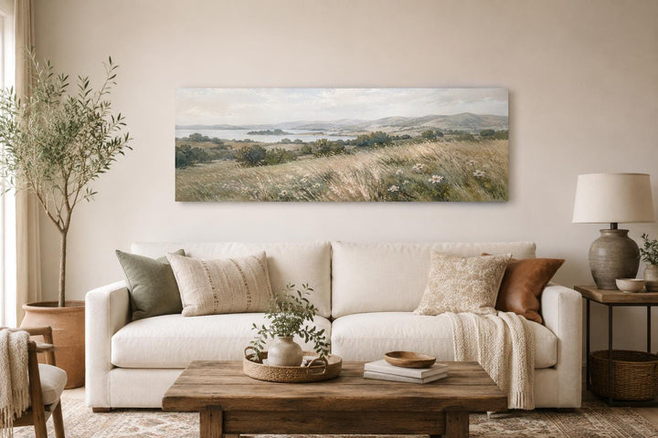Long horizontal canvas art of a neutral farmhouse grass field landscape above a white sofa.