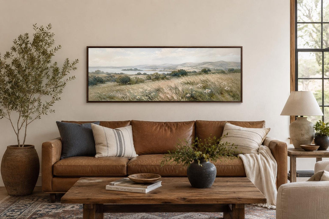 Long horizontal canvas of a farmhouse grass field landscape above a brown leather sofa.
