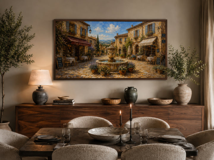 Framed canvas wall art of a sunlit French village street with a fountain above a dining sideboard.