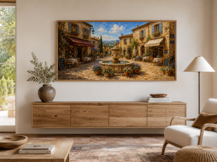 Framed canvas wall art of a sunlit French village street in Provence above a wooden sideboard.