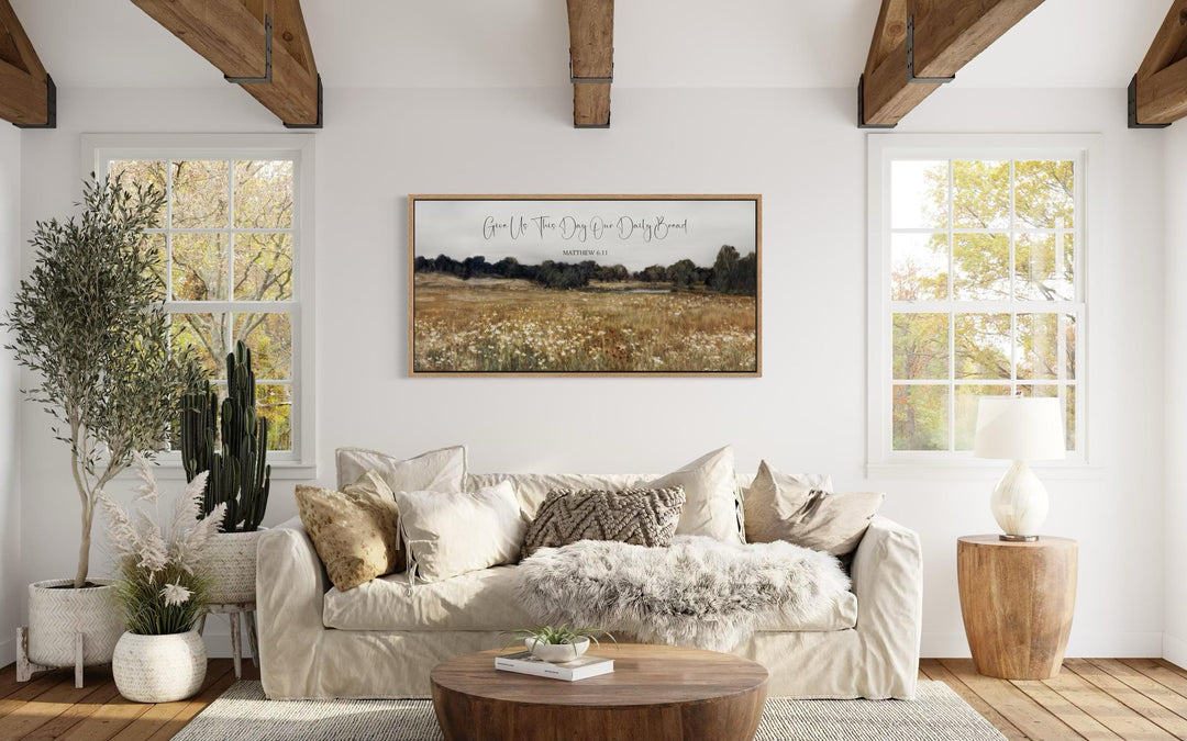 Framed landscape wall art with Matthew 6:11 scripture above a cozy, neutral-toned living room sofa.