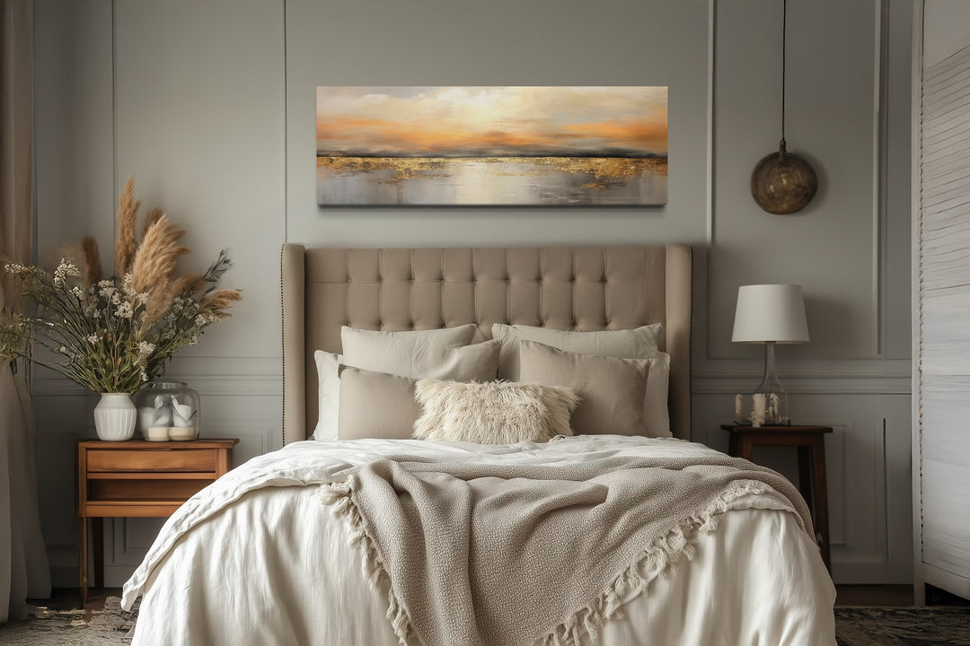 Gold and silver ocean sunset horizontal wall art hanging above a tufted beige headboard in a bedroom.
