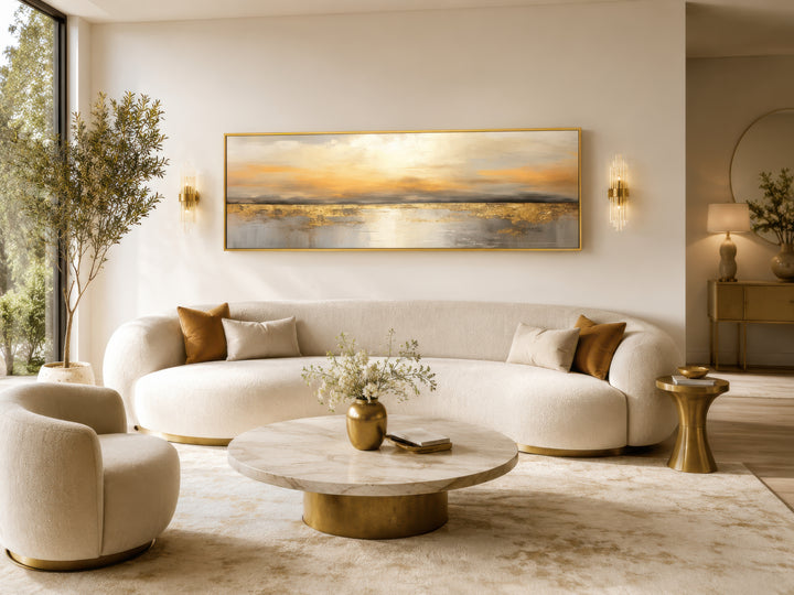 Long horizontal gold and silver abstract ocean sunset wall art hanging above a white curved sofa.