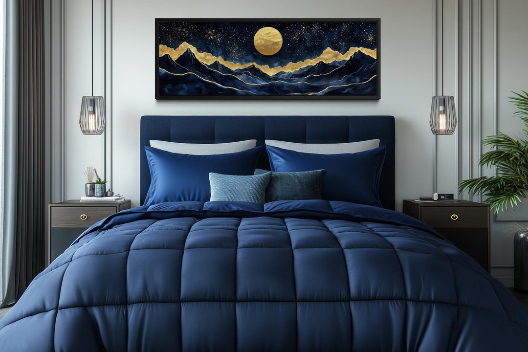 Panoramic gold and navy canvas print of a moonlit mountain landscape above a modern blue bed.