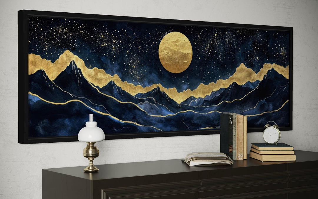 Panoramic canvas print of a gold moon and navy mountains in a black frame above a dark wood console.