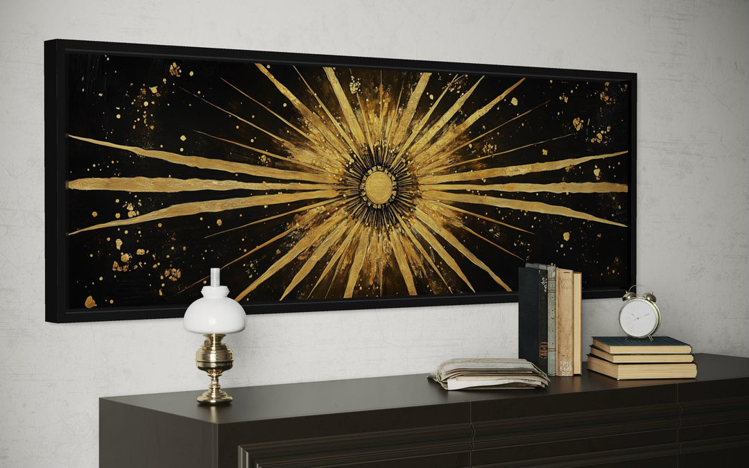 Long horizontal black and gold canvas painting of a radiant sunburst above a dark wood dresser.