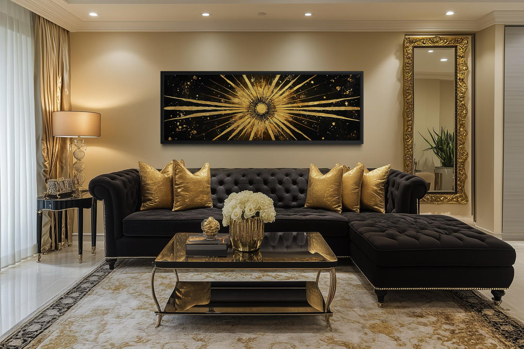A long horizontal black and gold canvas painting of a radiant sunburst above a black velvet sofa.