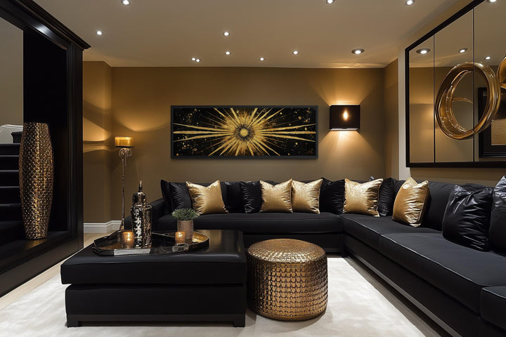 A long horizontal black and gold canvas painting of a radiant sunburst above a black sectional sofa.
