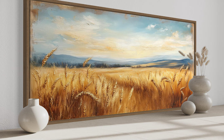 Golden wheat field farmhouse canvas wall art in a wooden frame above a neutral surface with vases.