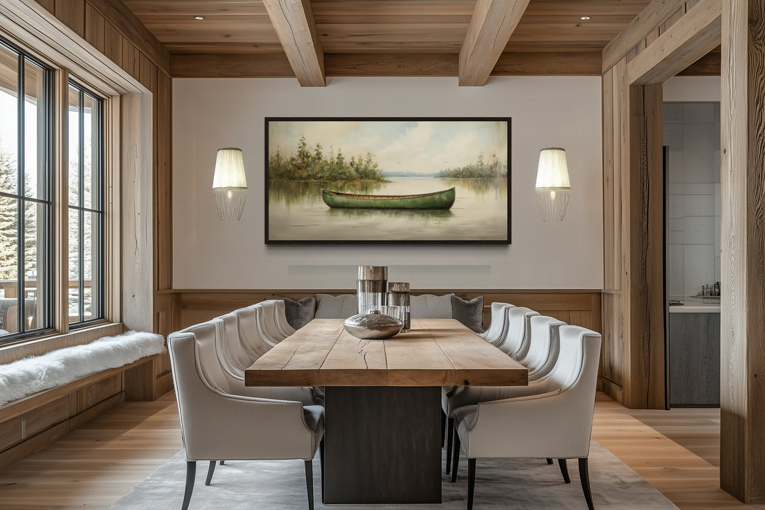 Framed canvas painting of a green canoe on a calm lake, hung in a rustic wood-paneled dining room.