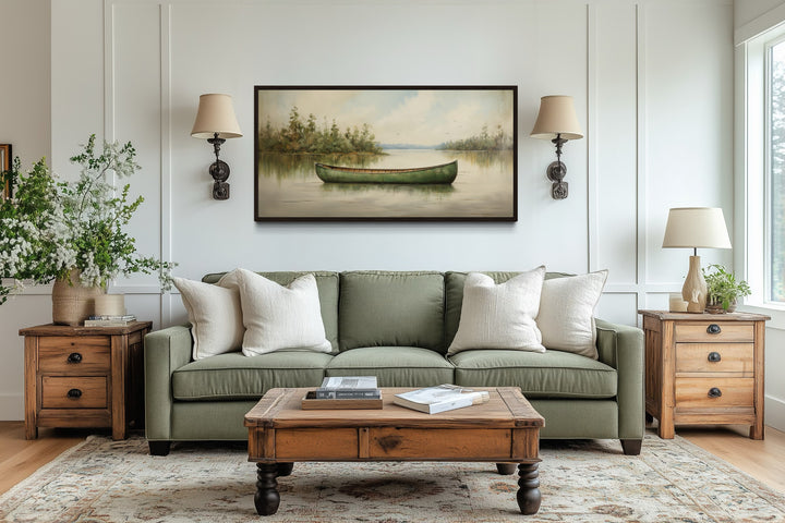 Framed canvas painting of a green canoe on a calm lake, styled above a green sofa in a rustic room.