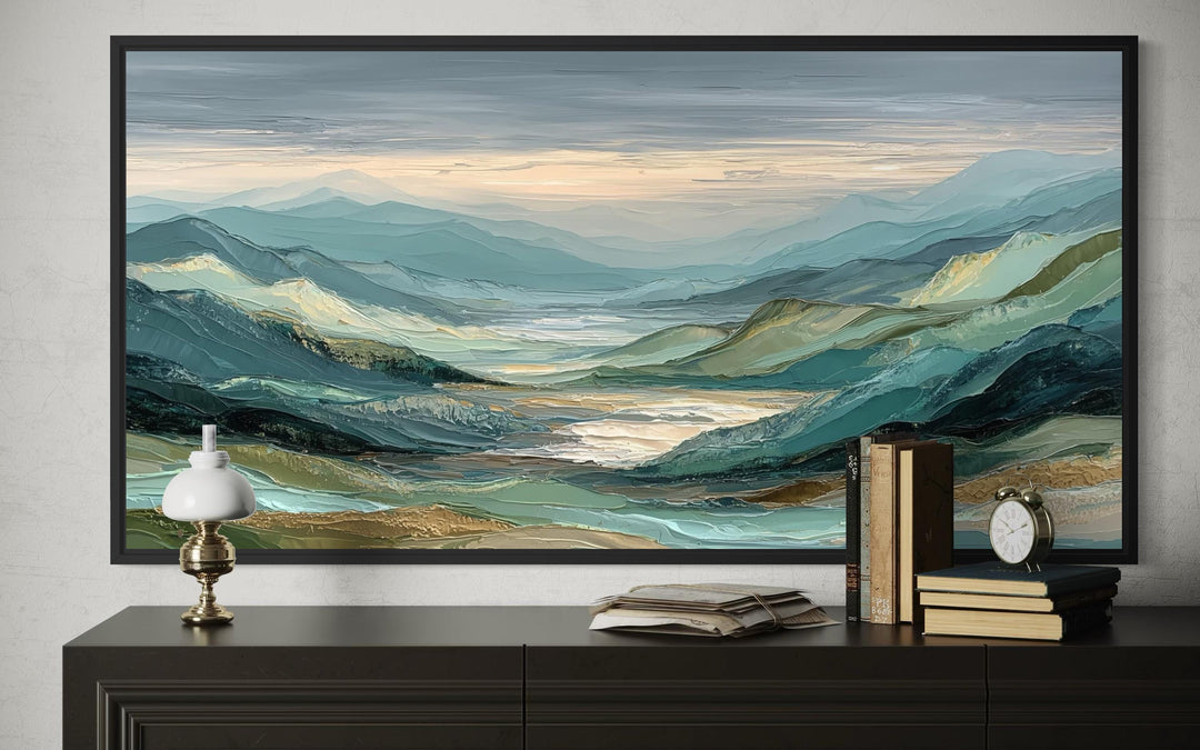 A framed horizontal painting of a textured green, grey, and gold mountain landscape on a dark console.