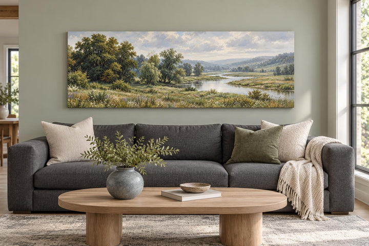 Long horizontal canvas wall art of a green valley and river landscape above a grey sofa.