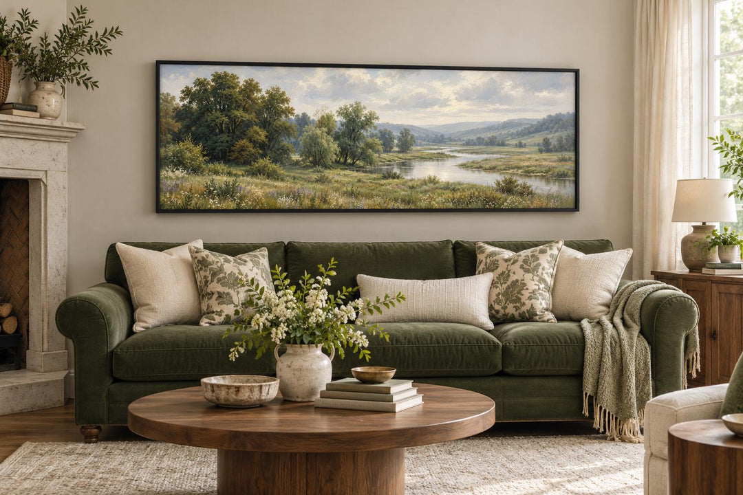 Long horizontal canvas wall art of a green valley and river landscape above a green sofa.