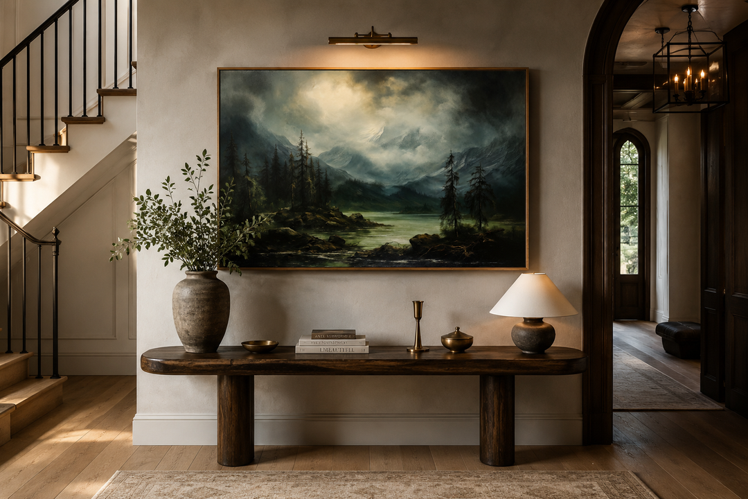 Moody vintage landscape painting of a misty lake and mountains in a gold frame above a wood console.