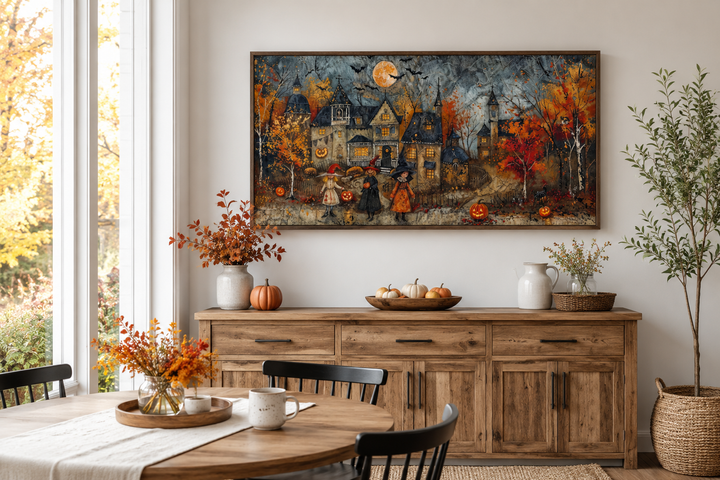 Framed canvas art of children trick-or-treating in a whimsical village above a wooden sideboard.
