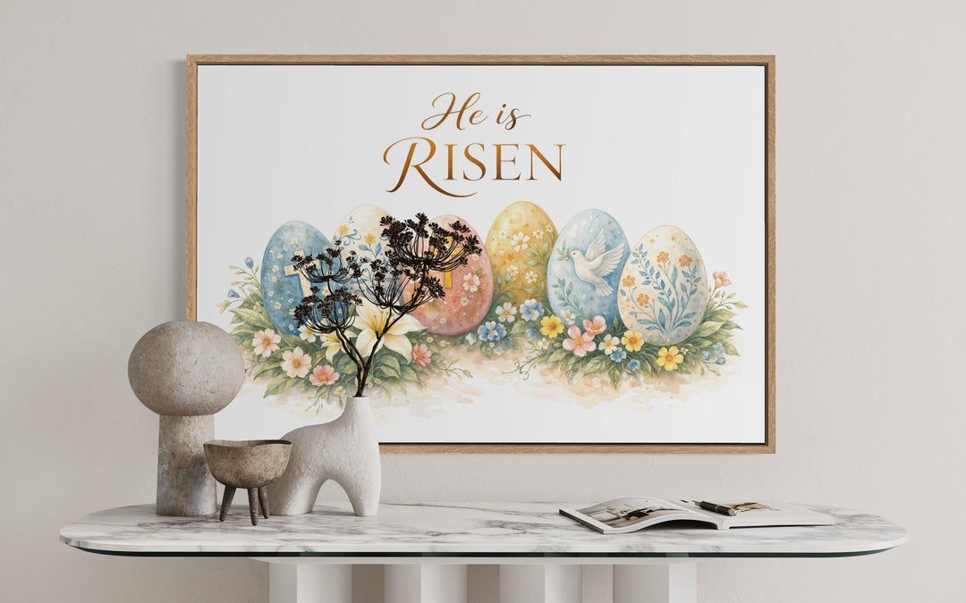 He Is Risen wall art featuring watercolor Easter eggs and flowers in a wooden frame on a marble table.
