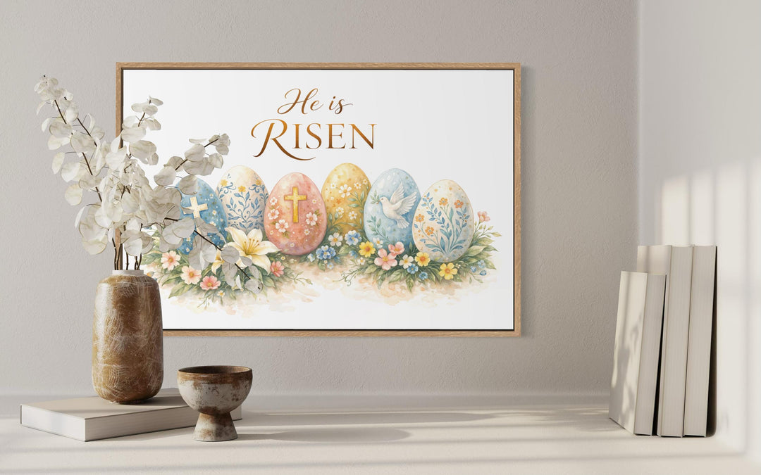 A framed canvas print featuring Easter eggs and the text He is Risen displayed on a neutral wall.