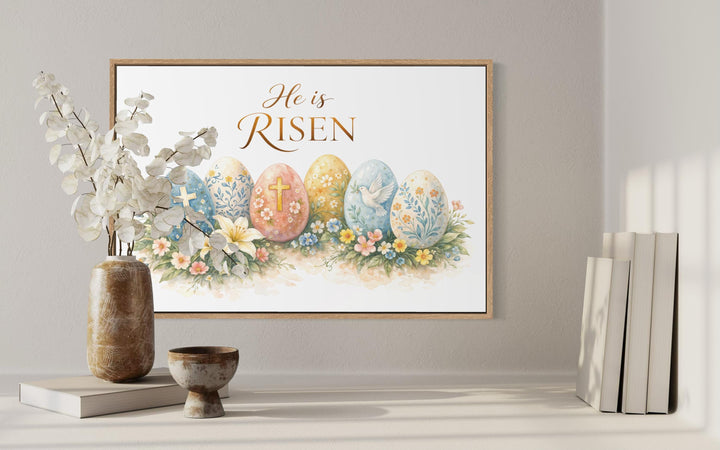 A framed canvas print featuring Easter eggs and the text He is Risen displayed on a neutral wall.
