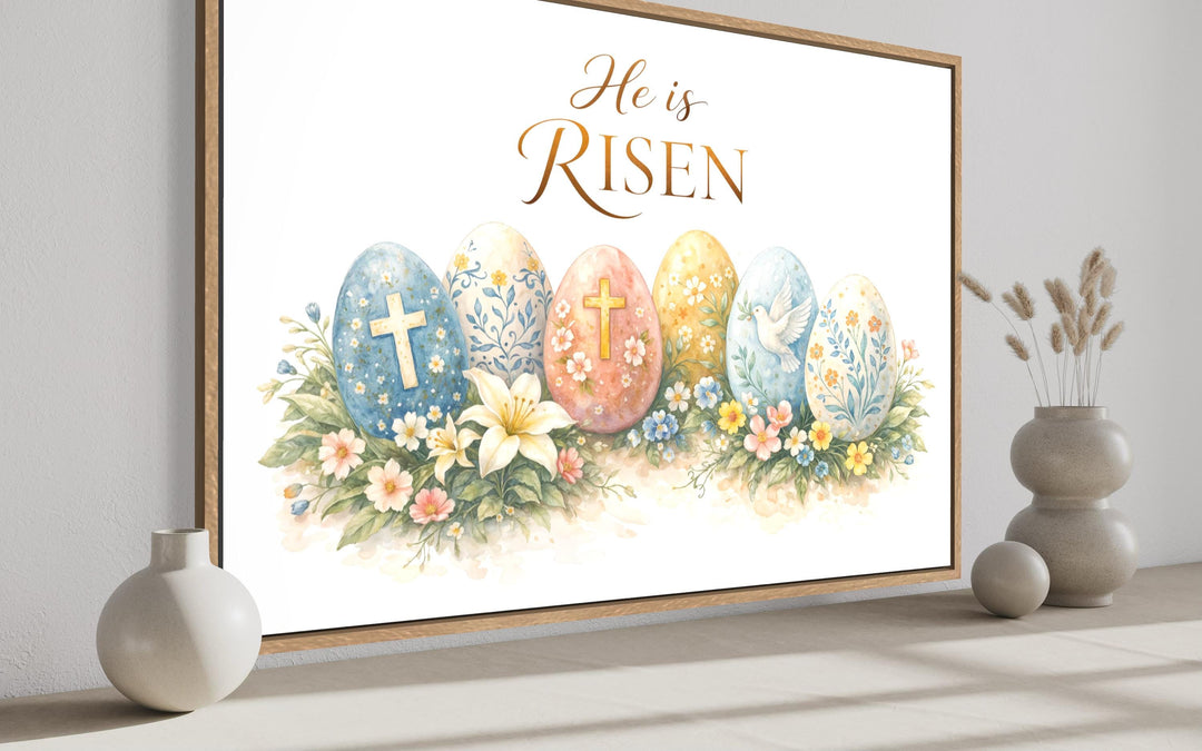 He Is Risen wall art featuring watercolor Easter eggs, crosses, and flowers in a wooden frame.