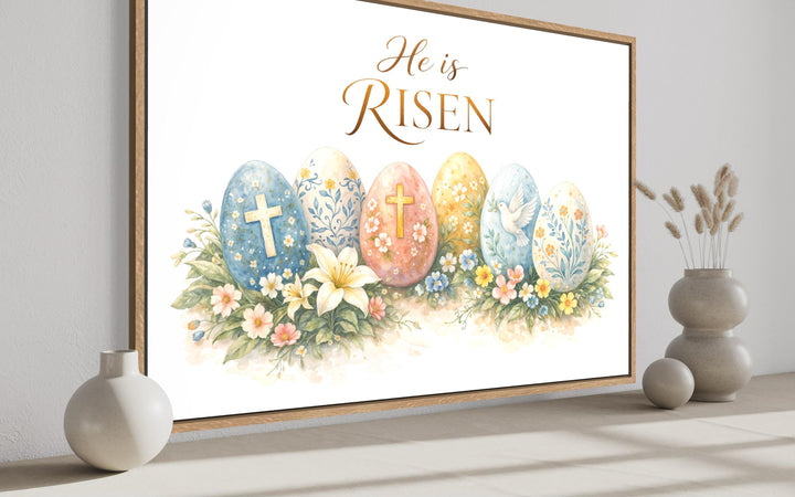 He Is Risen wall art featuring watercolor Easter eggs, crosses, and flowers in a wooden frame.