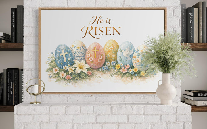 He Is Risen wall art featuring a cross and floral Easter eggs in a wooden frame on a white mantel.