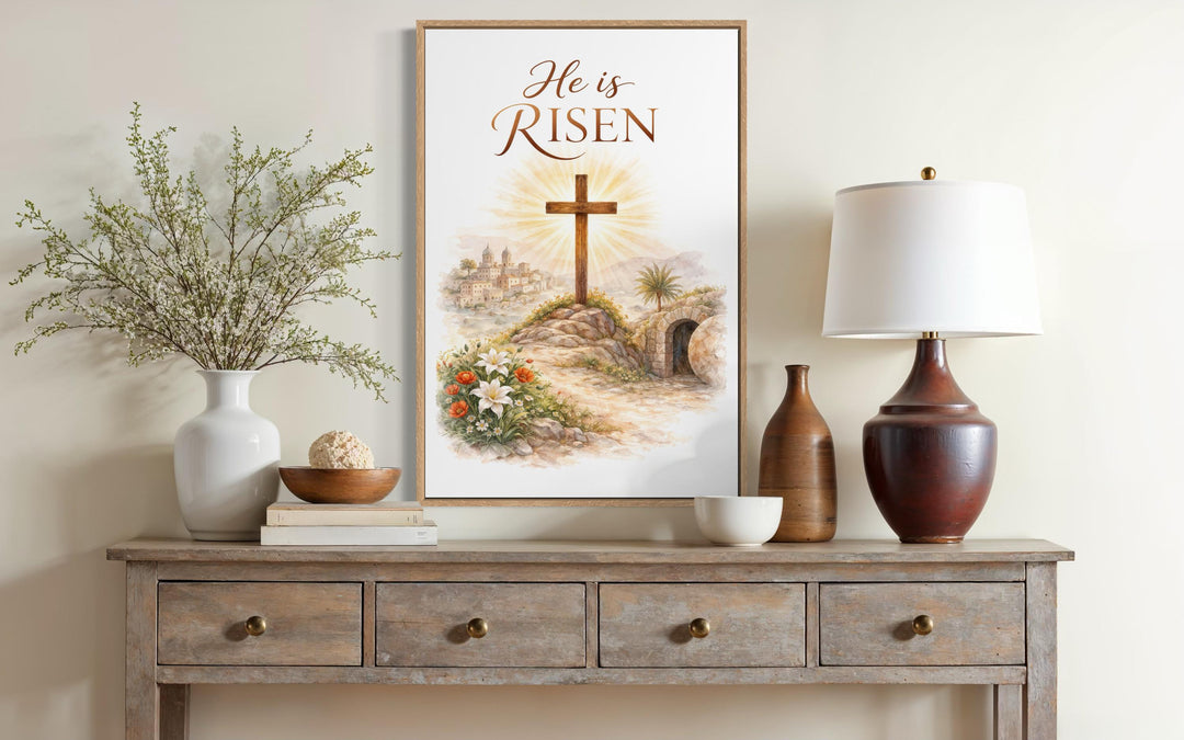 A framed He Is Risen canvas print featuring a cross and empty tomb on a rustic wooden console table.