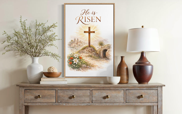 A framed He Is Risen canvas print featuring a cross and empty tomb on a rustic wooden console table.
