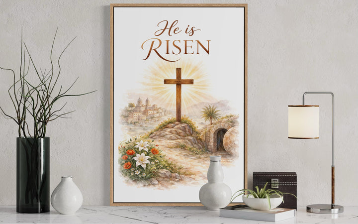 A framed watercolor print of a cross and empty tomb with the text He is RISEN on a white wall.