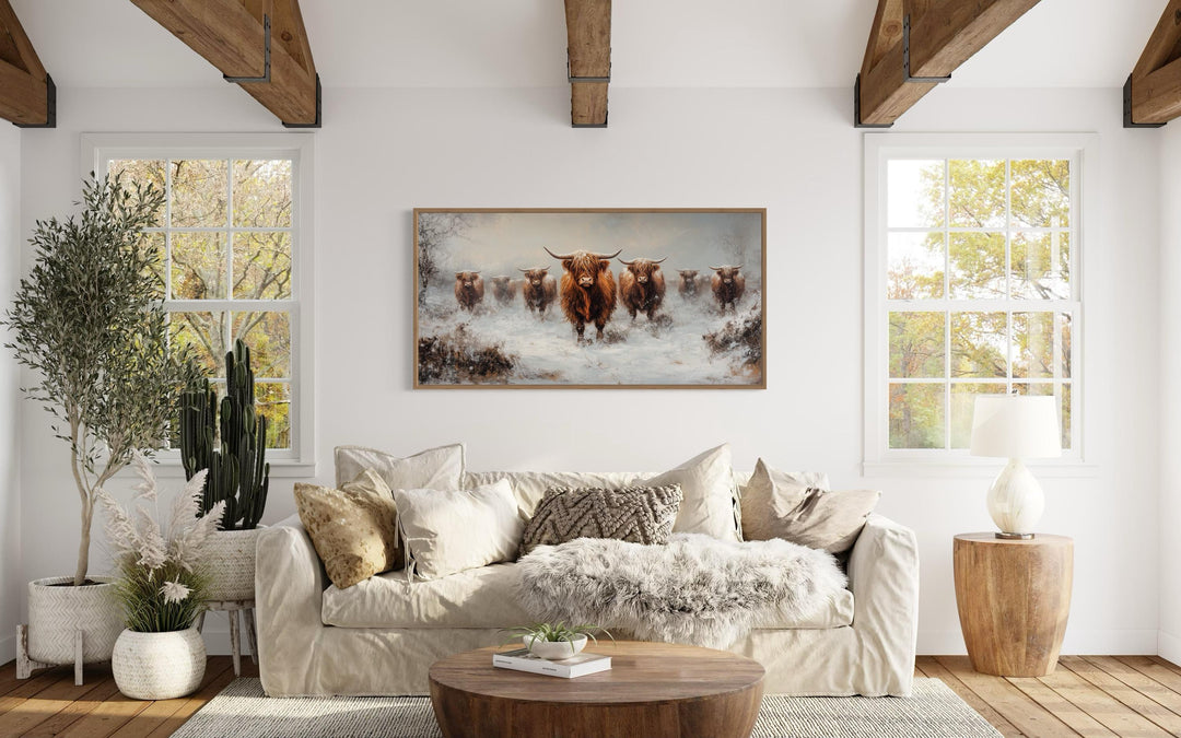 Framed canvas wall art of a herd of Highland cows in a snowy field above a couch in a rustic room.