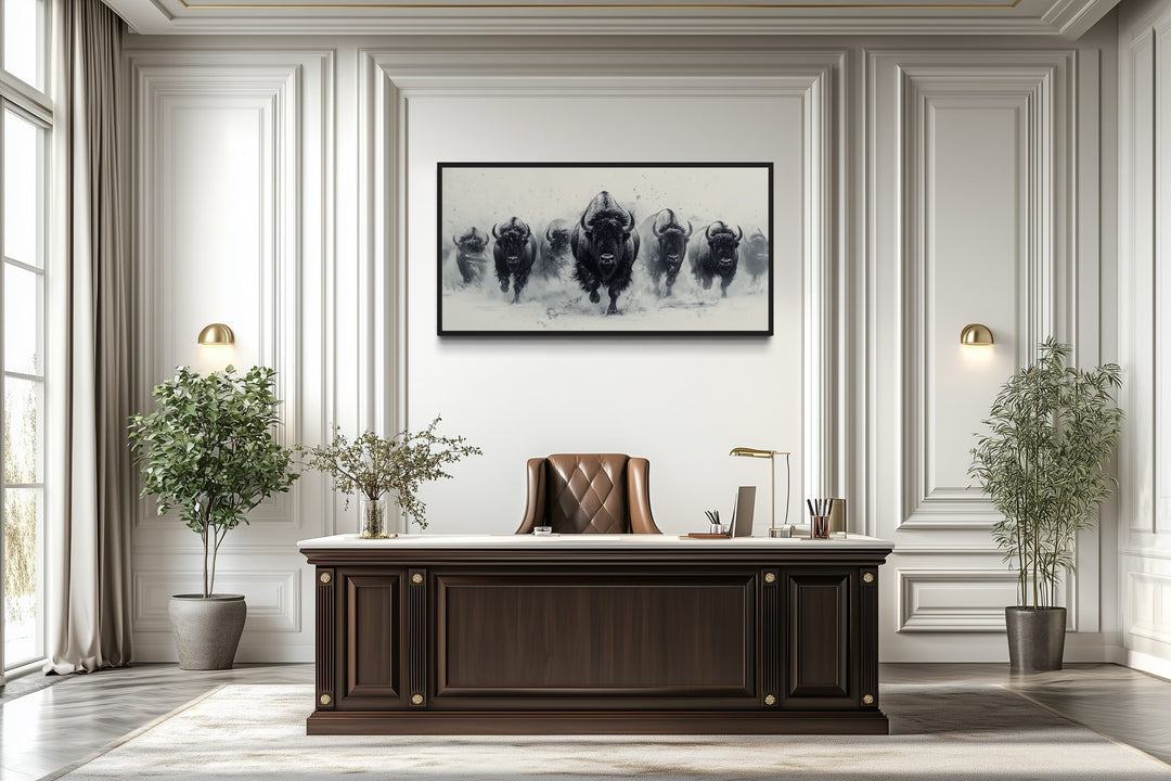 Black and white framed canvas wall art of a bison herd running in snow above a dark wood desk.