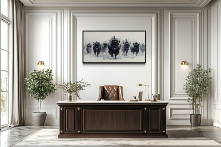 Black and white framed canvas wall art of a bison herd running in snow above a dark wood desk.