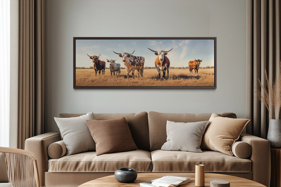 Panoramic framed canvas wall art of a Texas Longhorn cow herd in a field above a tan sofa.