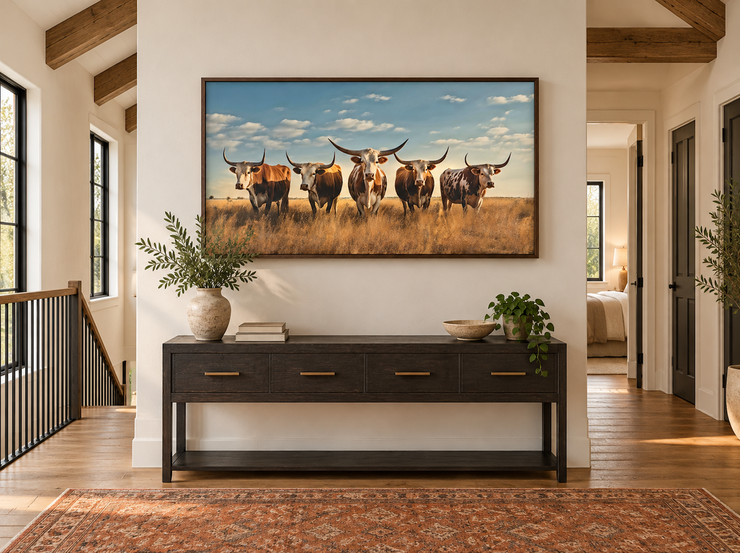 Framed horizontal canvas art of five Texas Longhorns in a sunlit field above a dark wood console.