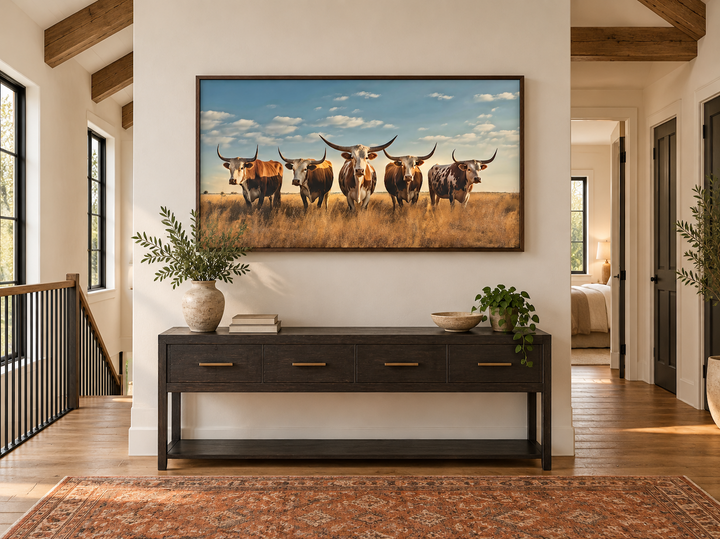 Framed horizontal canvas art of five Texas Longhorns in a sunlit field above a dark wood console.