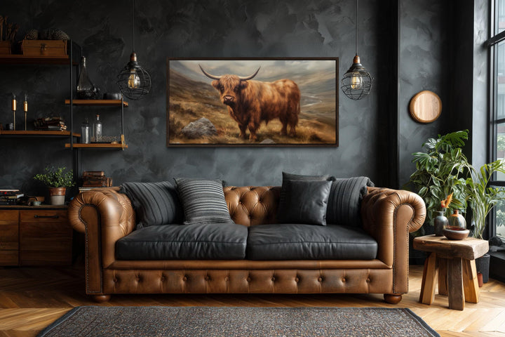 A framed canvas print of a rustic highland cow hangs above a leather sofa in a dark, moody room.
