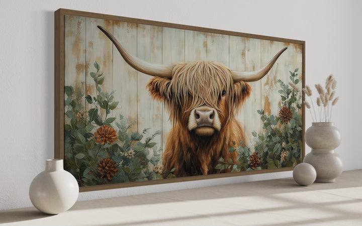 Highland cow framed canvas wall art with flowers on a distressed wood background in a rustic room.