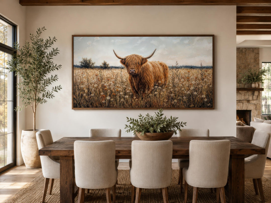 Highland Cow in Wildflower Meadow canvas print in a rustic frame above a wooden dining table.