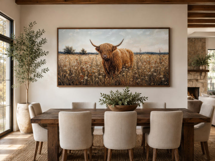 Highland Cow in Wildflower Meadow canvas print in a rustic frame above a wooden dining table.