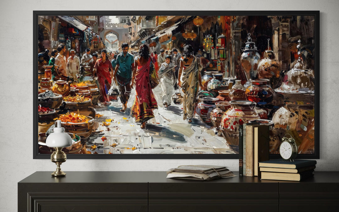 Framed Indian market painting on a dark sideboard with books, a lamp, and a clock.