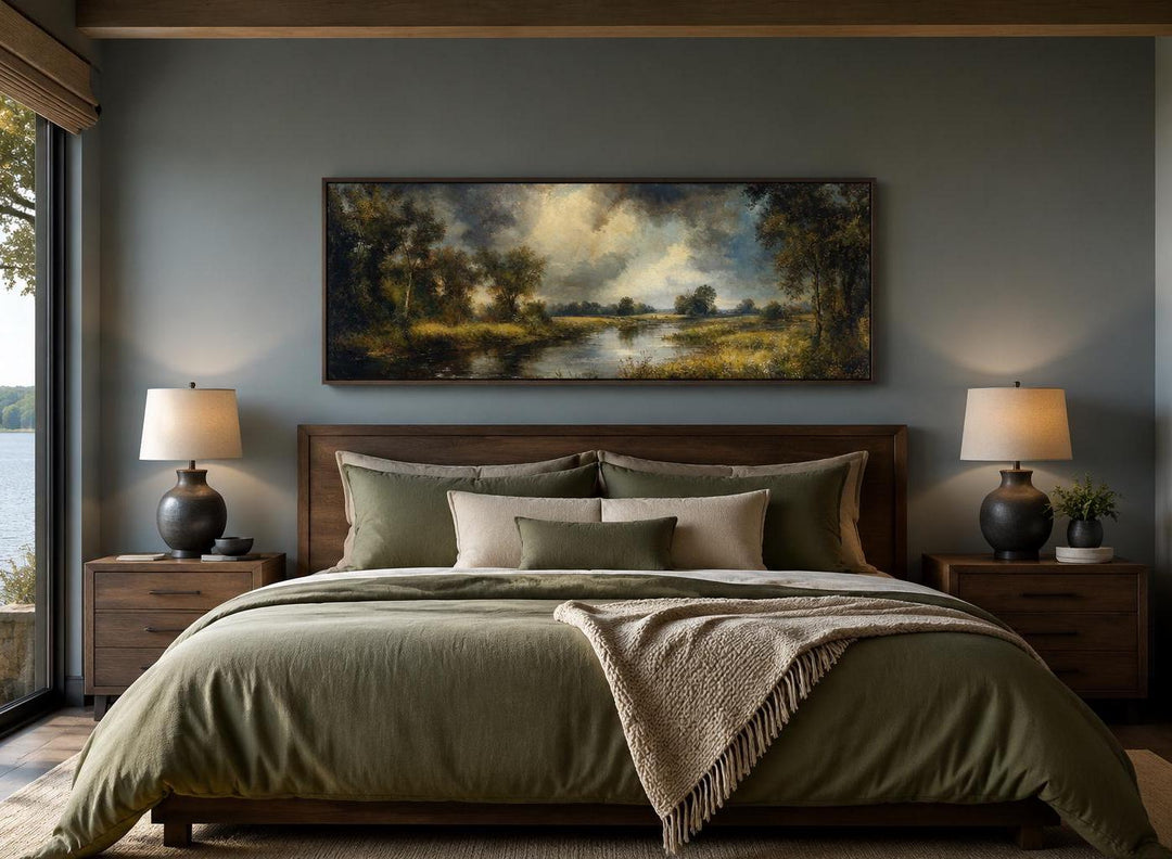 Vintage oil painting of a lake forest landscape in a dark wood frame above a bed.