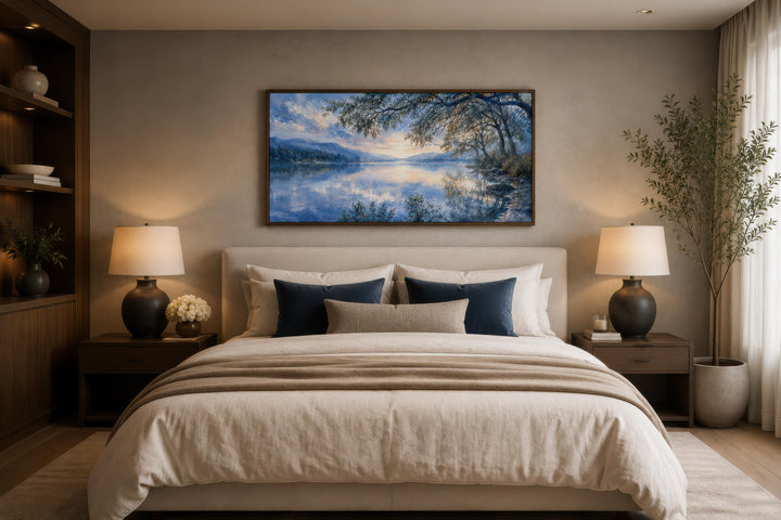 Framed horizontal canvas print of a blue lake landscape painting above a bed in a neutral bedroom.