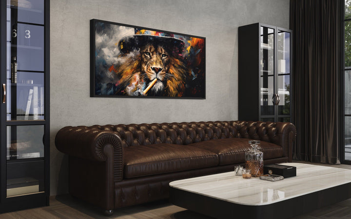 Framed canvas print of a lion smoking a cigar hanging above a brown leather Chesterfield sofa.