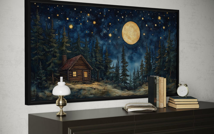 Log Cabin in the Moonlit Woods canvas wall art displayed in a black frame above a dark dresser.