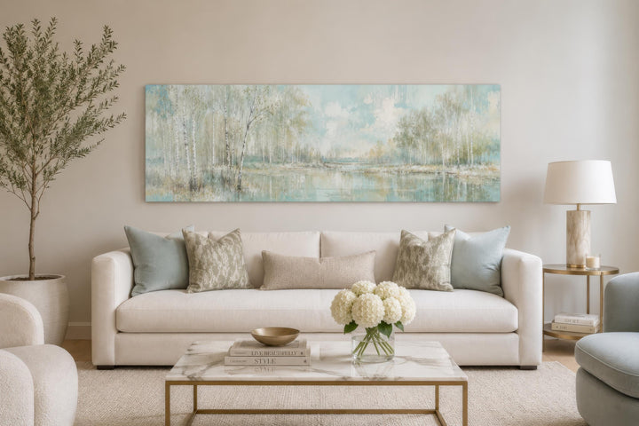 Long horizontal canvas print of a misty birch tree forest over a sofa in a neutral living room.