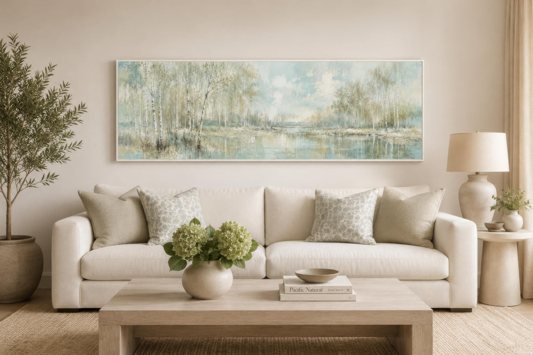 Long horizontal canvas print of a misty birch tree forest over a sofa in a neutral living room.